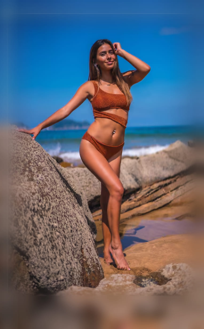 A woman standing barefoot on the rocks at the water's edge in a rust bikini, one hand in her hair, with the surf and a headland behind her